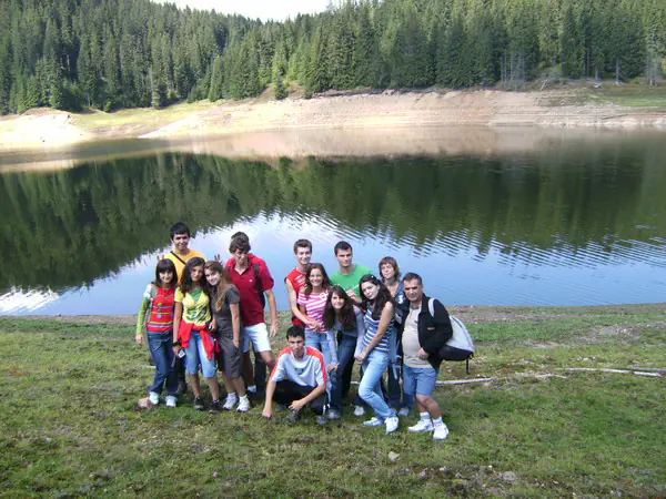 Grup de adolescenți pozând pe malul lacului, cu pădurea reflectată în apă