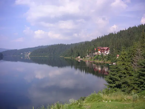 Lacul Beliș–Fântânele, liniștit, oglindind norii, cu o pensiune pe malul împădurit