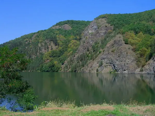 Versant stâncos și împădurit reflectat în apa verde a Lacului Beliș–Fântânele