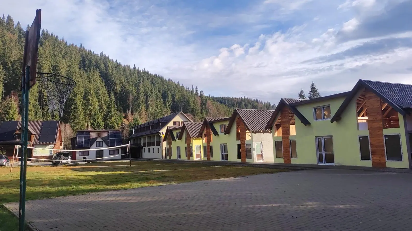 Șir de case moderne galbene, cu frontoane din lemn, lângă o curte pavată și pădurea de brazi