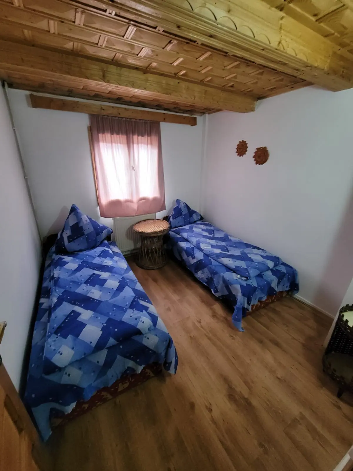 Cameră cu două paturi cu cuverturi albastre, pereți albi și tavan cu grinzi de lemn